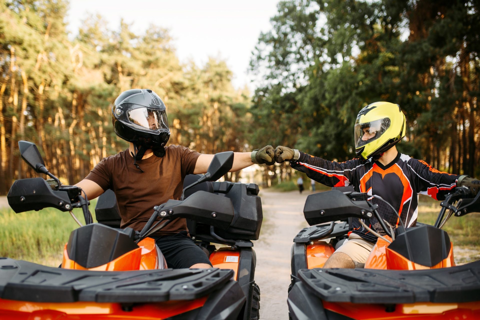 Two people on ATVs fist-bump in a forest, wearing helmets and protective gear, showcasing outdoor adventure and camaraderie.
