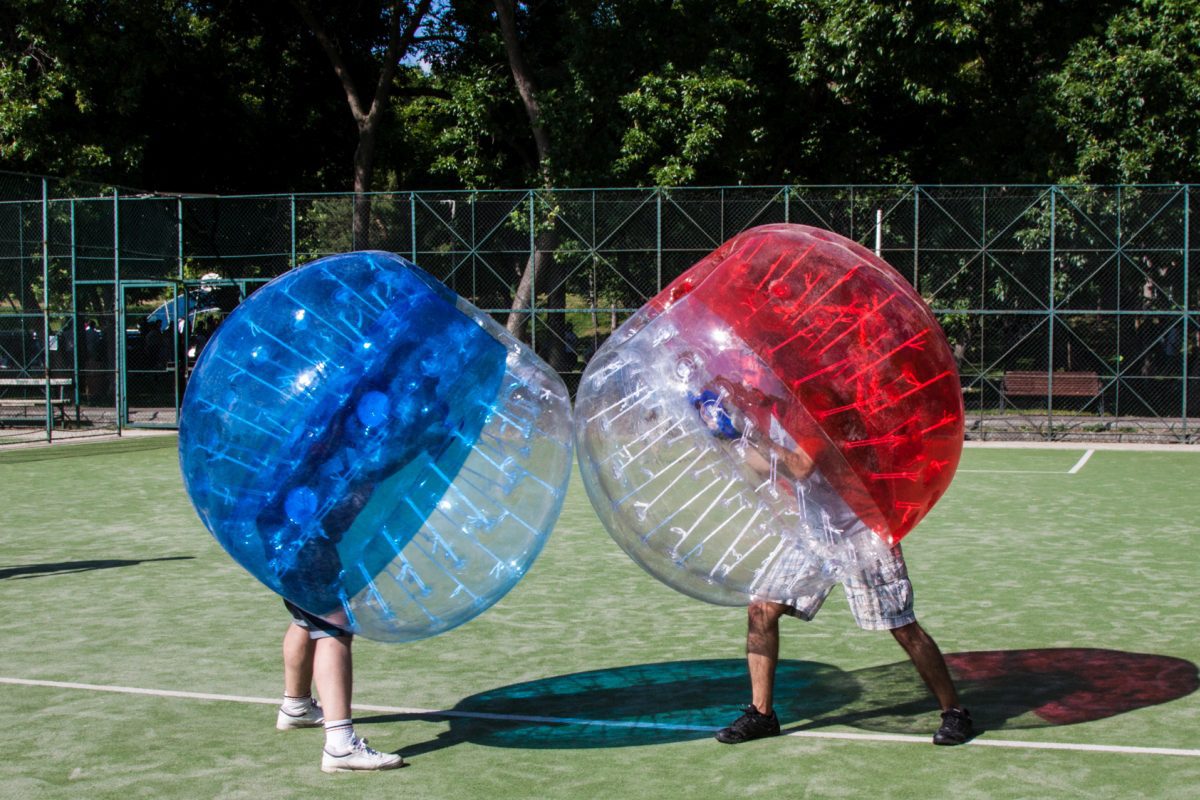 Two people playing bubble soccer on a green field, wearing blue and red inflatable suits.