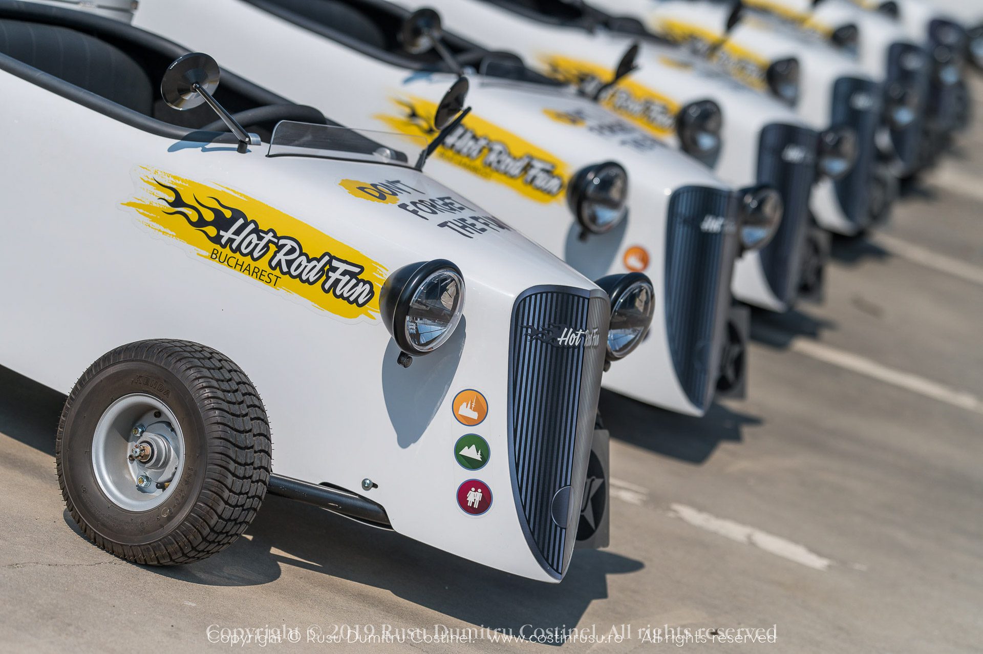 Row of white hot rod cars with Hot Rod Fun Bucharest logo parked in a line on sunny pavement.