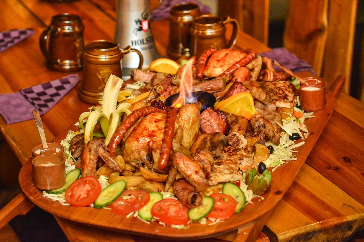Assorted grilled meats and vegetables on a platter with sauces and garnish, set on a wooden table with rustic mugs.