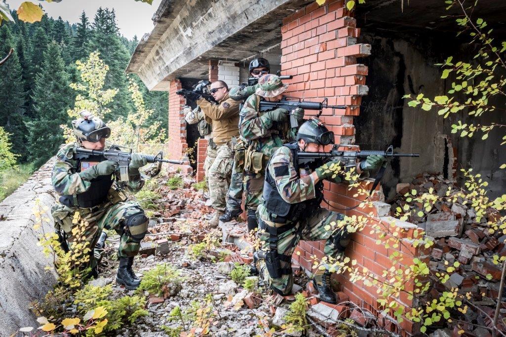 Soldiers in camouflage gear move tactically through a forested, brick building during a military training exercise.