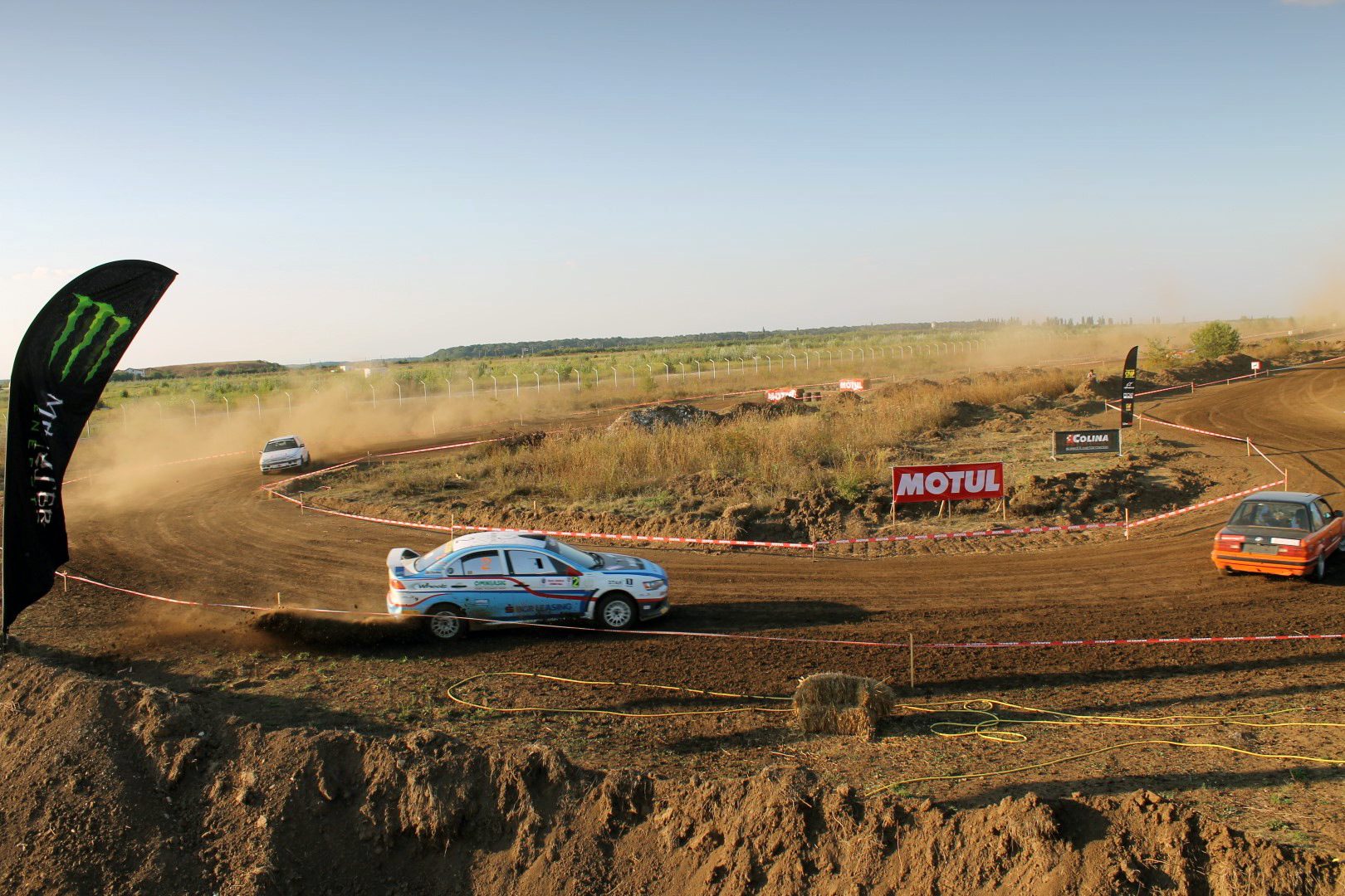 Rally cars racing on a dirt track, kicking up dust, surrounded by banners under a clear sky.