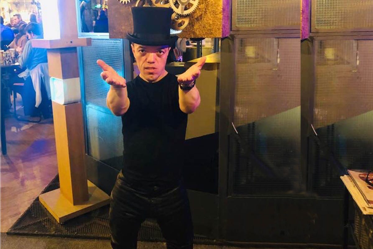 Man in top hat performing dramatic gesture in lively bar setting.