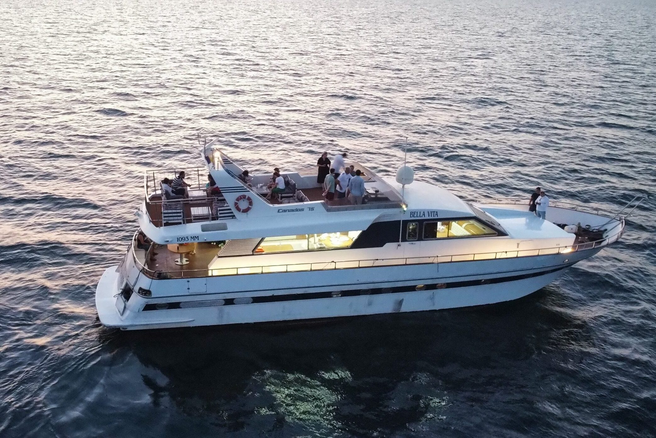 A yacht named Bella Vita cruises on calm waters at sunset with people onboard enjoying the view.
