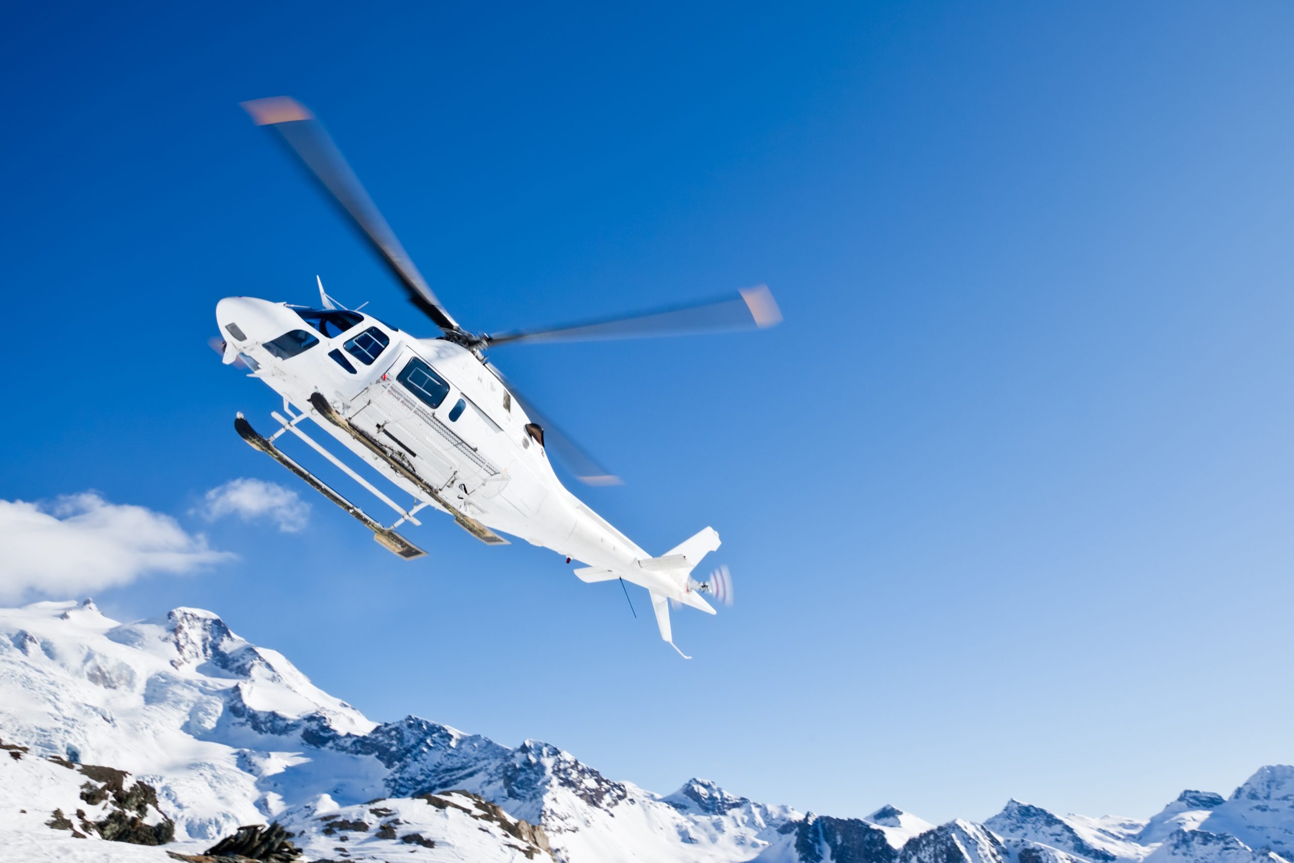 White helicopter flying over snowy mountains under clear blue sky.