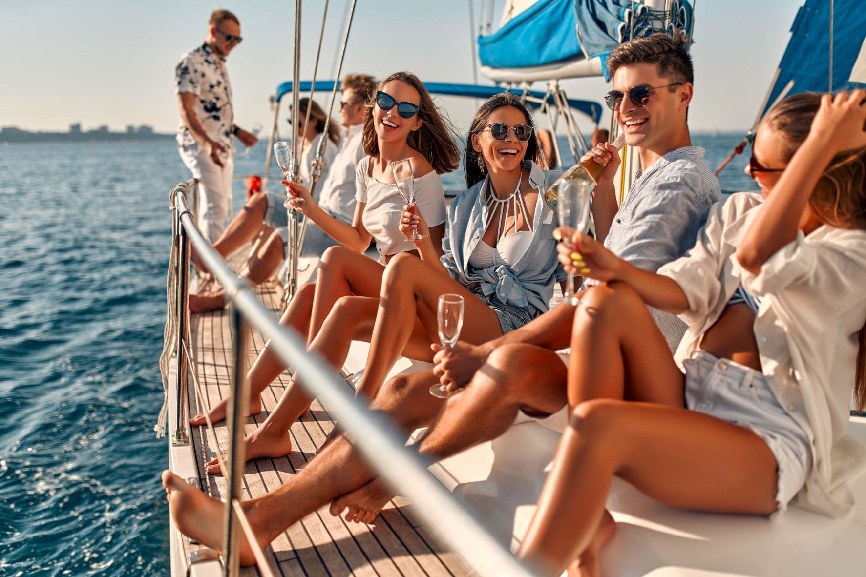 Friends enjoying a sunny day on a sailboat, smiling and holding champagne glasses, with the sea in the background.