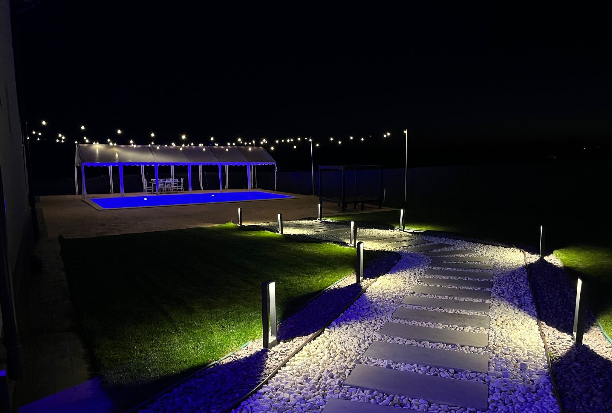 Pathway to a pool area illuminated by string lights and ground lamps, creating a serene nighttime ambiance.