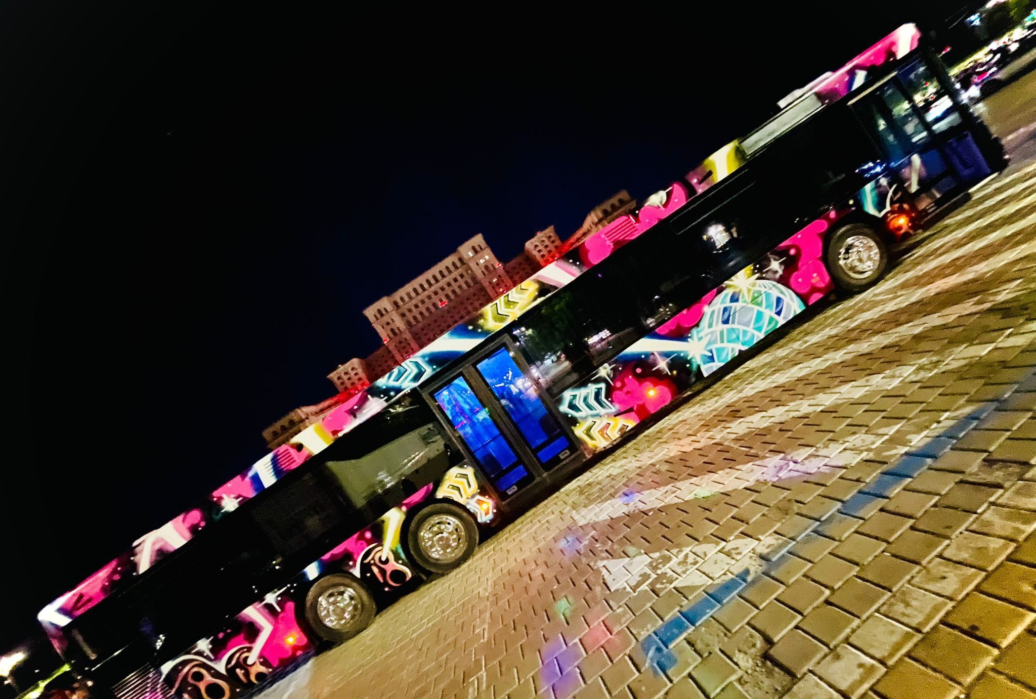 Colorful graffiti-adorned bus parked on brick pavement at night, vibrant lights and patterns visible.