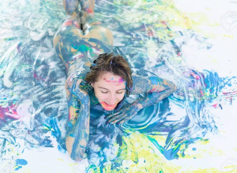 Top view of a naked woman in a spray of paint. A girl without clothes lies on a white floor in colored spots. Original body painting