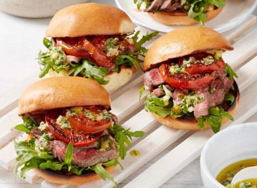 Gourmet steak sandwiches with arugula, grilled tomatoes, and herb topping on a wooden tray. Delicious lunch option.