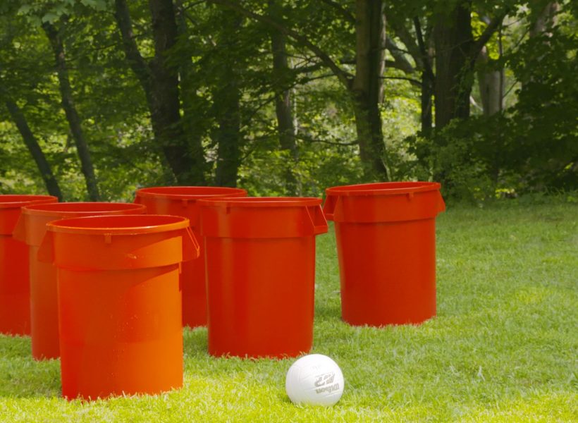 Six large orange buckets and a ball set up on green grass for an outdoor game in a wooded park setting.