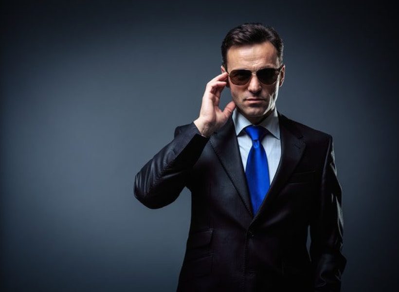 Man in dark suit and blue tie adjusting sunglasses against a gray background, exuding a confident and professional aura.