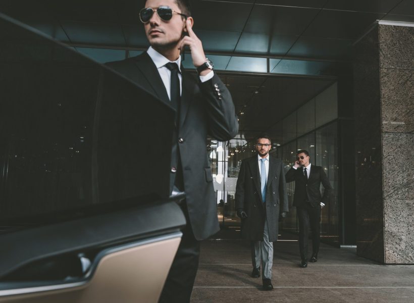 Men in suits and sunglasses outside a modern building, one speaking on a phone, conveying corporate security.