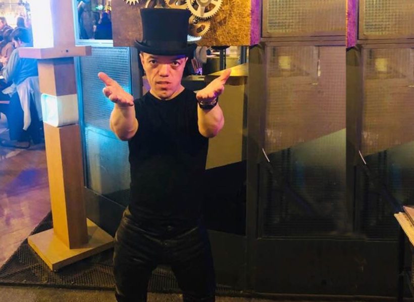Man in a top hat with open arms in a lively restaurant setting.
