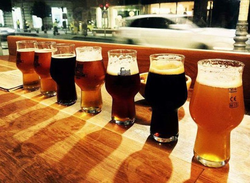 Romanian Beer Tasting 3 Seven craft beer glasses on a wooden table in a pub, city street view in the background.