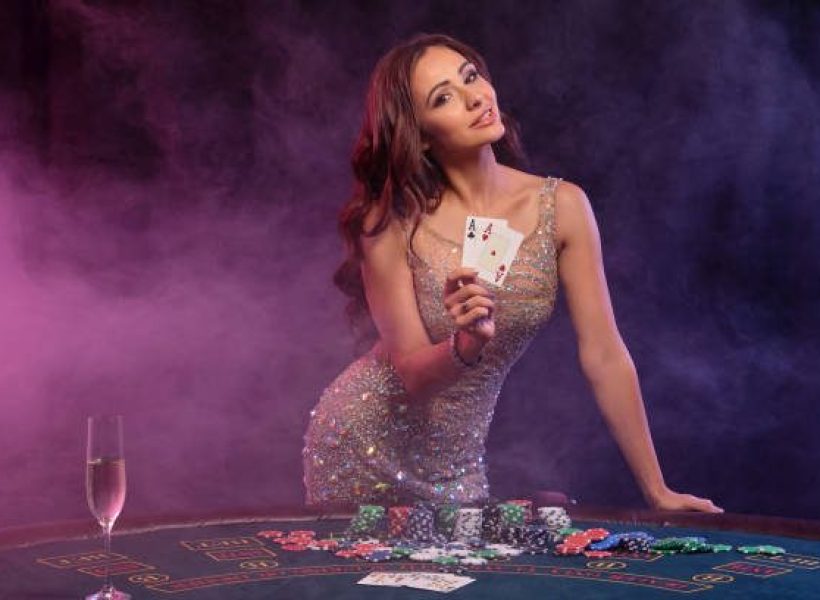 Pretty lady playing poker at casino. She showing cards posing at table with stacks of chips, money, champagne on it. Black, smoke background, colorful backlights. Gambling entertainment. Close-up.