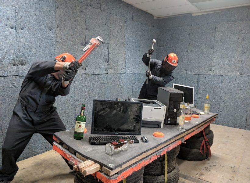 Two people in safety gear smashing electronics in a rage room, surrounded by empty liquor bottles, using tools.