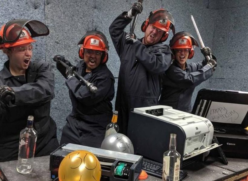 Group in safety gear smashing electronics with bats and tools in a controlled environment, expressing excitement.