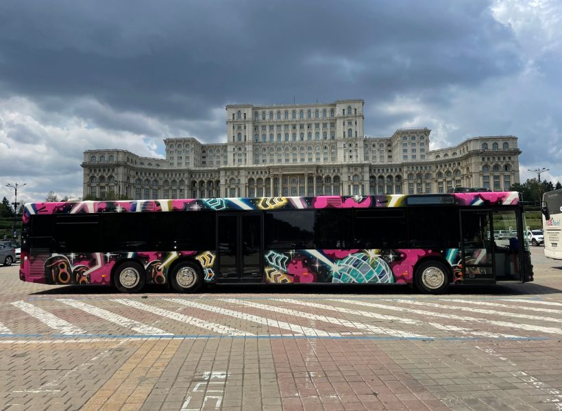 XXL Party Bus (2) Colorful bus with graffiti art parked in front of the grand Palace of the Parliament in Bucharest under a cloudy sky.