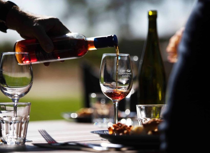atdw-AU0029576-632943fa314b83997c5c6dca-2000×1500 Pouring red wine into a glass at an outdoor table setting, with a bottle and sunlight in the background.