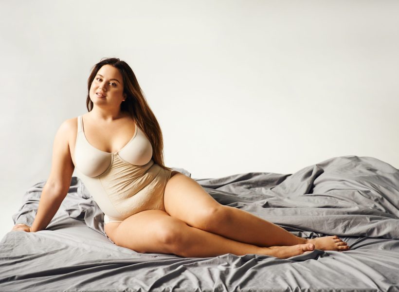 charming woman with natural makeup and plus size body wearing beige bodysuit and posing on bed with grey bedding, body positive, figure type, bare feet, looking at camera