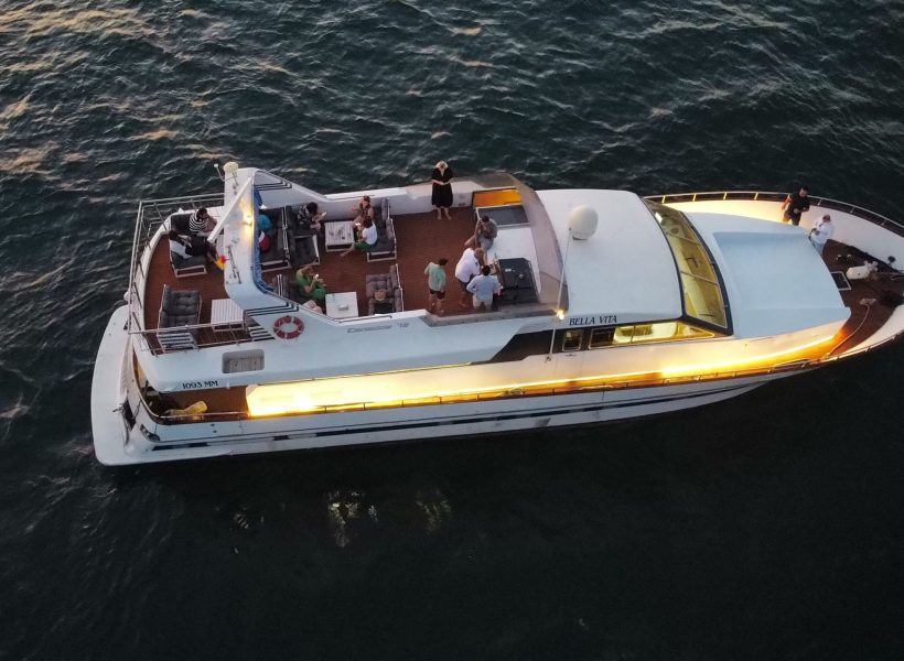 dji_fly_20230715_210916_646_1689444599755_photo_optimized-scaled Aerial view of a yacht named Bella Vita with people onboard, cruising on calm ocean waters at dusk.