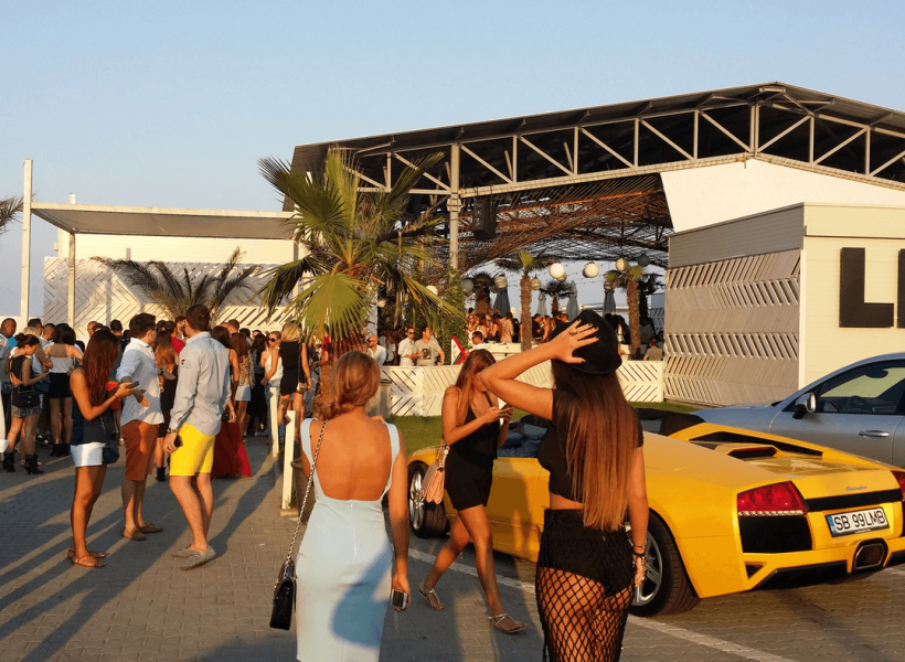 People gather at a vibrant outdoor event by a venue with stylish cars and palm trees in the background.