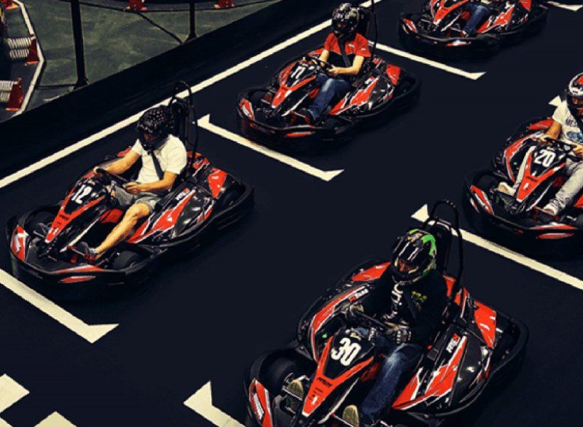 indoor-karting1 Drivers racing in go-karts on an indoor track, wearing helmets, engaging in competitive fun.