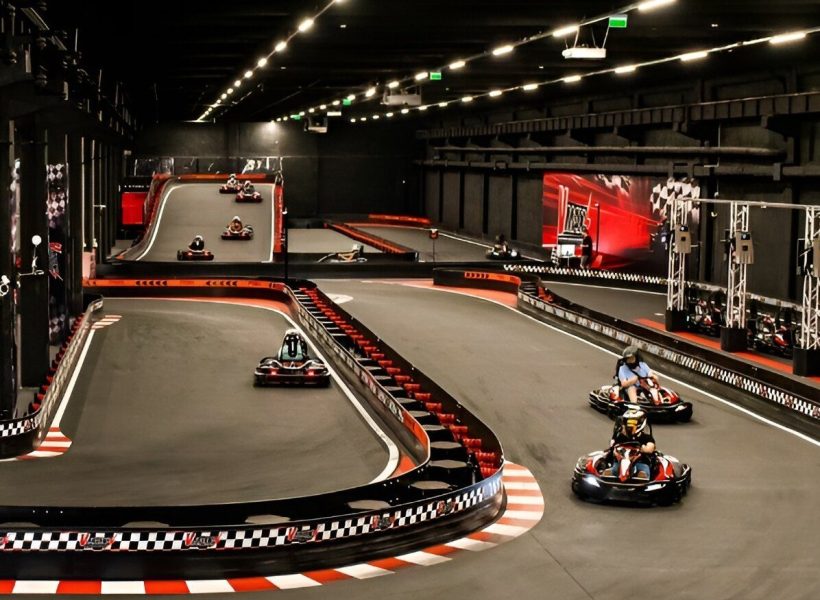 indoor-karting2 Indoor go-kart racing track with several karts in action, featuring dynamic curves and vibrant lighting.