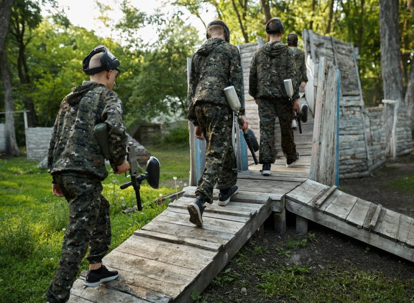 Paintball team shoots with guns, warriors in camouflages on playground in the forest. Extreme sport with pneumatic weapon and paint bullets or markers, active military game outdoors, combat tactics