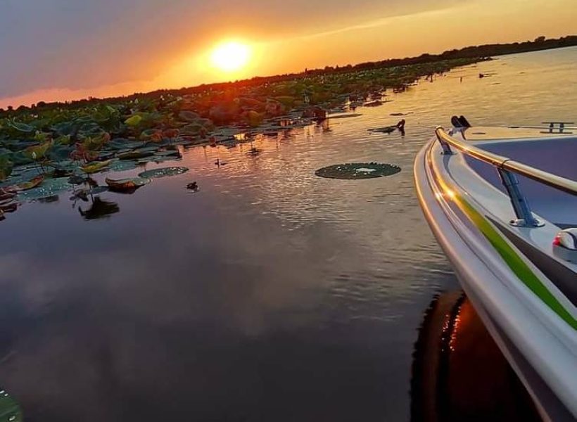 Sunset over a serene lake with lilypads and a boat in view, reflecting vibrant colors on the water.
