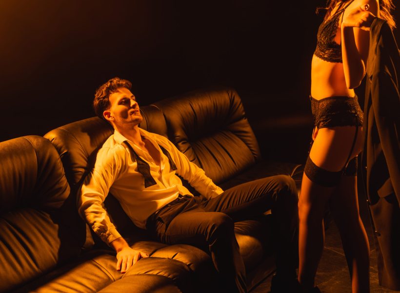 seductive woman in underwear holding jacket and standing near man on black Man in open dress shirt on couch, woman in lingerie stands near, dimly lit elegant setting.