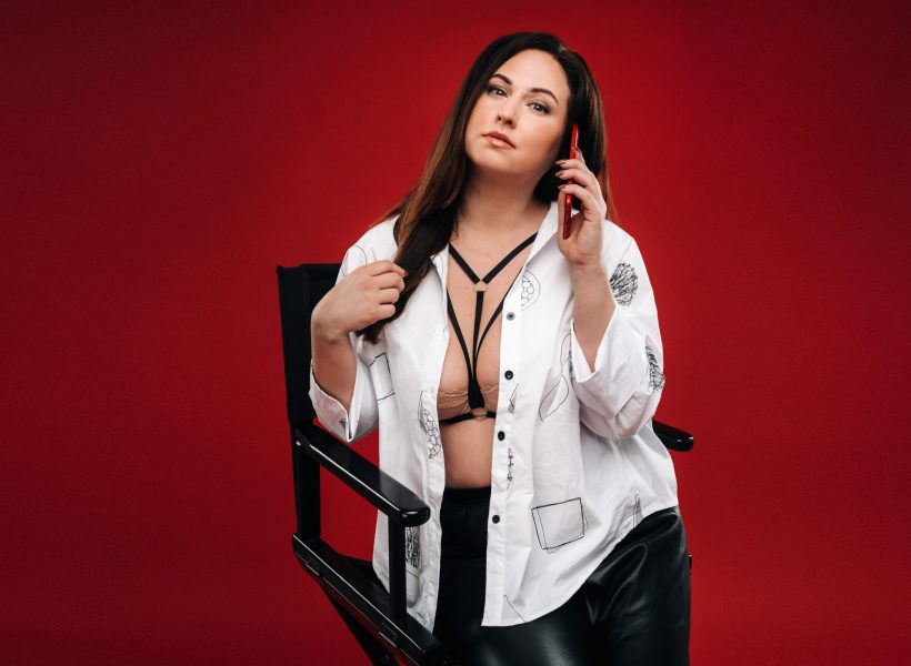 Woman in stylish white blouse and leather pants sitting on a chair with a red phone, set against a red background.