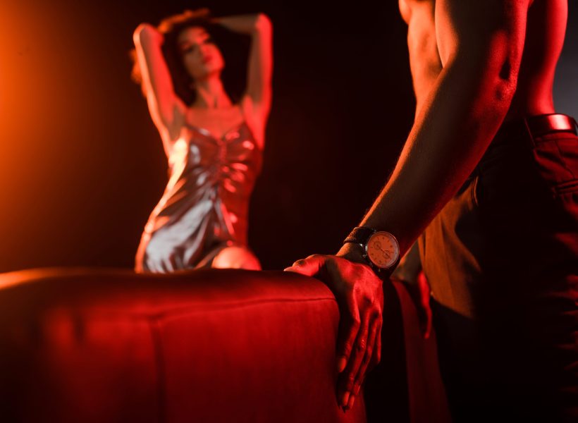 shirtless african american man standing near couch and seductive woman on blurred background Dimly lit fashion scene featuring elegant couple, focus on man's wristwatch, orange lighting creates a dramatic ambiance.